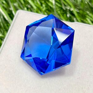 May include: A faceted, brilliant blue gemstone with a geometric shape. The gem is displayed on a white surface, with green grass visible in the background. The gem's facets reflect light, creating a sparkling effect.