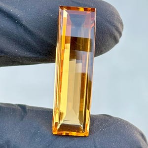 Puede incluir: Una gema rectangular y facetada con un cálido tono ámbar-naranja. El corte de la gema muestra reflejos internos y una superficie pulida. La gema se sostiene sobre un fondo oscuro, resaltando su claridad y color. Esta gema se usa en joyería.