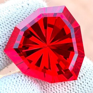 May include: A faceted, vibrant red gemstone with a complex, geometric design. The gem's edges are sharply defined, and the interior features a starburst pattern. The gemstone is held against a white knitted background.