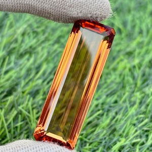 May include: A rectangular, faceted gemstone with a warm orange and amber color gradient. The gem is held by a gloved hand against a backdrop of green grass. The cut of the gem is a long, slender rectangle with angled edges.