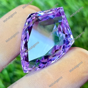 Könnte beinhalten: Ein facettierter, hellvioletter Edelstein mit einem komplexen, geometrischen Schliff. Der Edelstein wird zwischen Fingern gehalten, mit einem grünen Hintergrund. Die Facetten des Edelsteins reflektieren das Licht und erzeugen einen funkelnden Effekt. Der Text "Mehakgems" wiederholt sich.