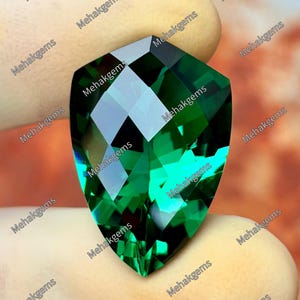 May include: A shield-shaped, faceted gemstone with a deep, vibrant emerald green colour. The stone features a complex geometric cut with multiple reflective surfaces that create light and dark patterns. The text Mehakgems is repeated.