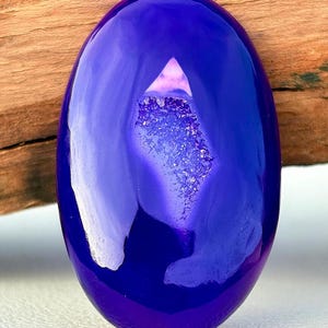 Può includere: Una gemma ovale lucidata di un profondo colore indaco. La pietra ha una superficie riflettente e una cavità piena di formazioni cristalline scintillanti. La gemma è posta su uno sfondo di legno naturale.