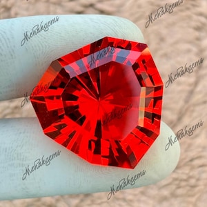 Op de afbeelding: Een gefacetteerde, achthoekige, helderrode edelsteen. De edelsteen heeft een complexe interne structuur met vele reflecterende oppervlakken, waardoor een briljant lichtspel ontstaat. De edelsteen wordt vastgehouden door een gehandschoende hand, met het woord "Mehakgems" als watermerk.