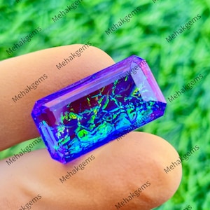 May include: A rectangular faceted gemstone with a deep purple base color and an internal pattern of iridescent green and blue flashes. The stone has sharp, clean edges and a polished surface that reflects light, creating a vibrant, multi-colored effect.