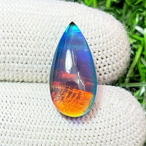 May include: A pear-shaped gemstone displaying a colour gradient. The top is teal, fading to blue, purple, and orange at the base. The stone has a reflective, polished surface, and is likely for jewellery.