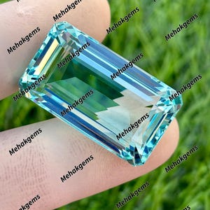 May include: A rectangular, aquamarine-colored gemstone with a faceted cut. The gem is held between two fingers against a blurred green background. The text "Mehakgems" is repeated around the gem.