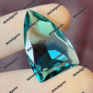 May include: A teal-coloured, faceted gemstone with an unusual, angular form. The gem is held against a neutral background, highlighting its clarity and colour. The text "Mehakgems" is repeated around the gem.