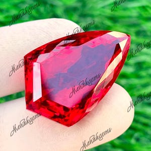 May include: A vibrant, deep red gemstone with a distinctive, elongated form. The gem is displayed against a blurred green backdrop, emphasizing its brilliant cut and intense colour. The facets of the gem reflect light, producing a sparkling effect. The image is well-lit, highlighting the gem's clarity.