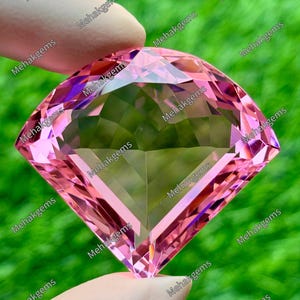 May include: A large, cushion-cut pink gemstone with a faceted surface that reflects light. The stone is transparent and displays a vibrant, uniform pink hue throughout its structure, showcasing precise geometric cuts and sharp, clean edges.