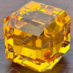 Könnte beinhalten: Ein facettierter, transparenter gelber Edelstein in Würfelform. Die Oberfläche des Steins weist mehrere geometrische Facetten auf, die das Licht reflektieren und ein helles, goldenes Aussehen erzeugen. Der Text Mehakgems ist auf dem Bild wiederholt.