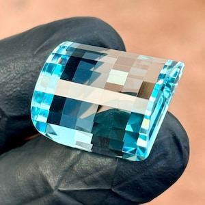 May include: A rectangular gemstone with a curved top, featuring a mosaic of light and dark blue hues. The facets create a reflective surface. The gem is held against a black surface, highlighting its colour and clarity.