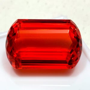 May include: A faceted, rectangular-cut gemstone in a vivid red colour. The gem has a polished surface with multiple facets, reflecting light. The shape is elongated with rounded ends, showcasing the gem's clarity and colour.