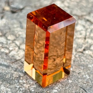 May include: A faceted, rectangular crystal in shades of amber and orange. The crystal has a square top and a vertical, columnar shape. The light refracts through the transparent material, creating a warm, glowing effect. The crystal is set on a rough, gray surface.