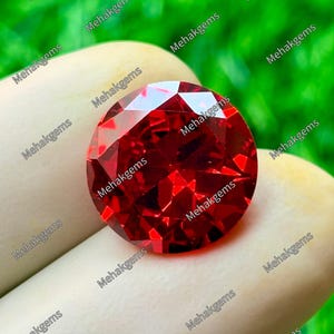 May include: A round, faceted, vibrant red gemstone with a brilliant cut. The stone displays multiple triangular and kite-shaped facets that reflect light, creating a sparkling appearance. The text Mehakgems is repeated across the image.