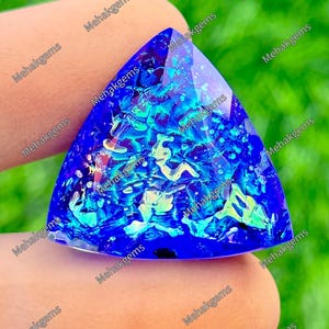 May include: A triangular-shaped gemstone featuring a deep blue base color with iridescent, multi-colored flecks of light blue, green, and yellow throughout the surface. The stone has a faceted top and is marked with the repeated text Mehakgems.