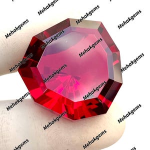 May include: A faceted, octagonal, deep red gemstone. The gem is held by a gloved hand. The gem has multiple facets that reflect light, creating a brilliant display. The text "Mehakgems" is repeated around the gem.