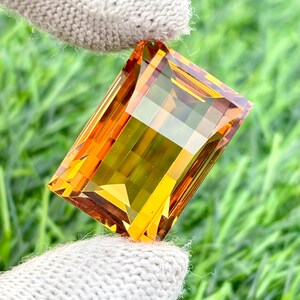 May include: A large, rectangular-cut gemstone with a vibrant blend of yellow, orange, and brown hues. The gem is held by a gloved hand, showcasing its facets and clarity. The background is a blurred green, suggesting a natural setting.