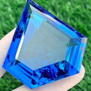 May include: A large, faceted gemstone in a vibrant blue colour. The gem is held against a backdrop of green grass, emphasising its clarity and brilliance. The cut of the stone creates multiple reflective surfaces.