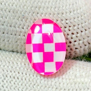 May include: An oval-shaped, pink and white checkered design. The pattern is a grid of alternating pink and white squares. The object has a glossy finish and is set against a textured, off-white background.