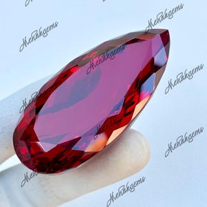 Könnte beinhalten: Ein facettierter, länglicher Edelstein in tiefroter Farbe. Der Stein wird von einer behandschuhten Hand gehalten, wodurch seine polierten Oberflächen und komplizierten Schnitte hervorgehoben werden. Die Form ähnelt einem Marquise-Schliff und reflektiert das Licht.