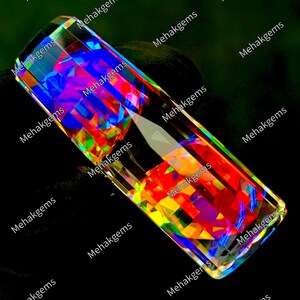 May include: A rectangular, faceted crystal prism displaying a vibrant spectrum of iridescent colors including red, yellow, and blue. The surface is highly reflective, creating geometric light patterns. The text Mehakgems is repeated across the image.