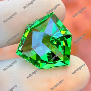 Puede incluir: Una gema de color verde brillante y vibrante con forma pentagonal y cortes geométricos multifacéticos intrincados que crean reflejos de luz nítidos. La piedra presenta una superficie pulida y está marcada con el texto repetido Mehakgems.
