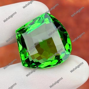 May include: A vibrant, faceted, cushion-cut green gemstone with a high-clarity, transparent appearance. The stone features multiple geometric facets that reflect light, creating a brilliant, sparkling effect across its surface. The text Mehakgems is repeated across the image.