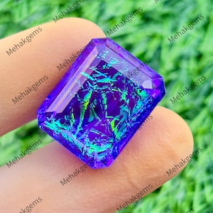 May include: A faceted rectangular gemstone with a deep purple base color and vibrant, iridescent green and blue internal inclusions that create a shimmering, splintered pattern throughout the stone's transparent, polished surface.