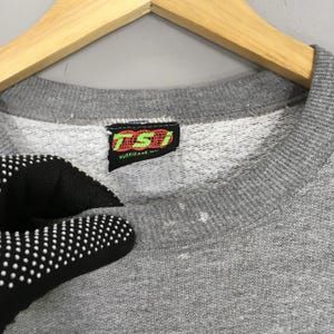 Puede incluir: Primer plano de una sudadera gris con cuello redondo. La etiqueta presenta las letras "TSI" en verde y naranja, con la palabra "HURRICANE" debajo. La sudadera est&aacute; colgada en una percha de madera.