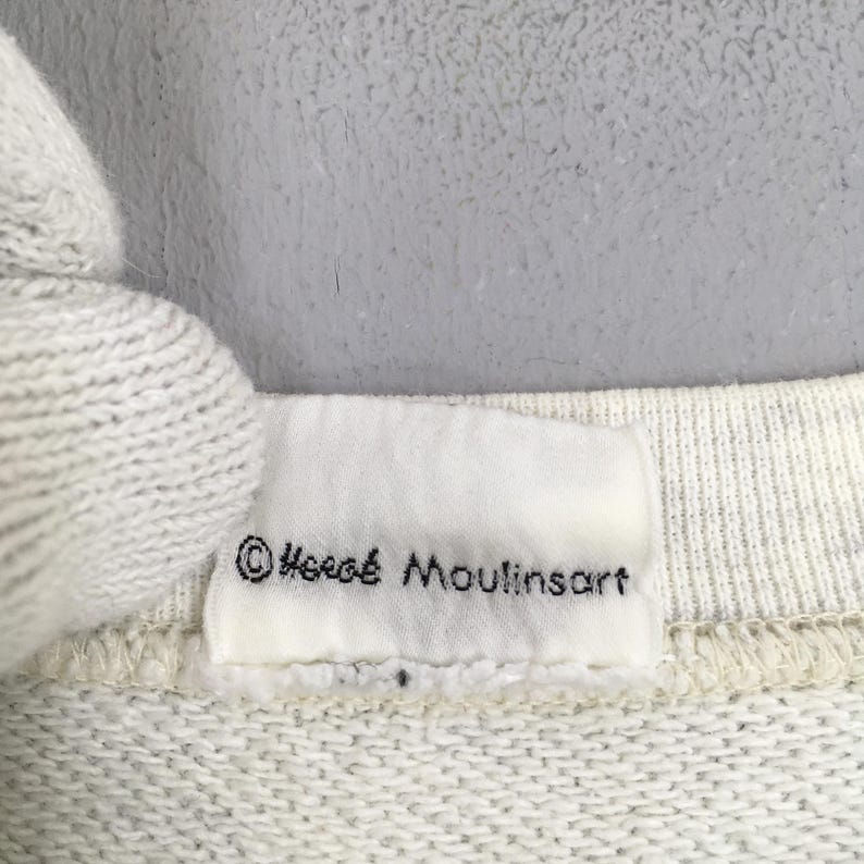 Peut inclure: Gros plan sur une &eacute;tiquette en tissu blanc avec le texte "&copy; Herg&eacute; Moulinsart" en noir. L'&eacute;tiquette est attach&eacute;e &agrave; un tissu clair et textur&eacute;. Le fond est gris clair.