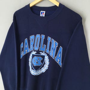 May include: A navy blue sweatshirt with the word "CAROLINA" in light blue block letters. Below the word is a light blue shield with a white wreath and the letters "NC". The sweatshirt has a crew neck and long sleeves.