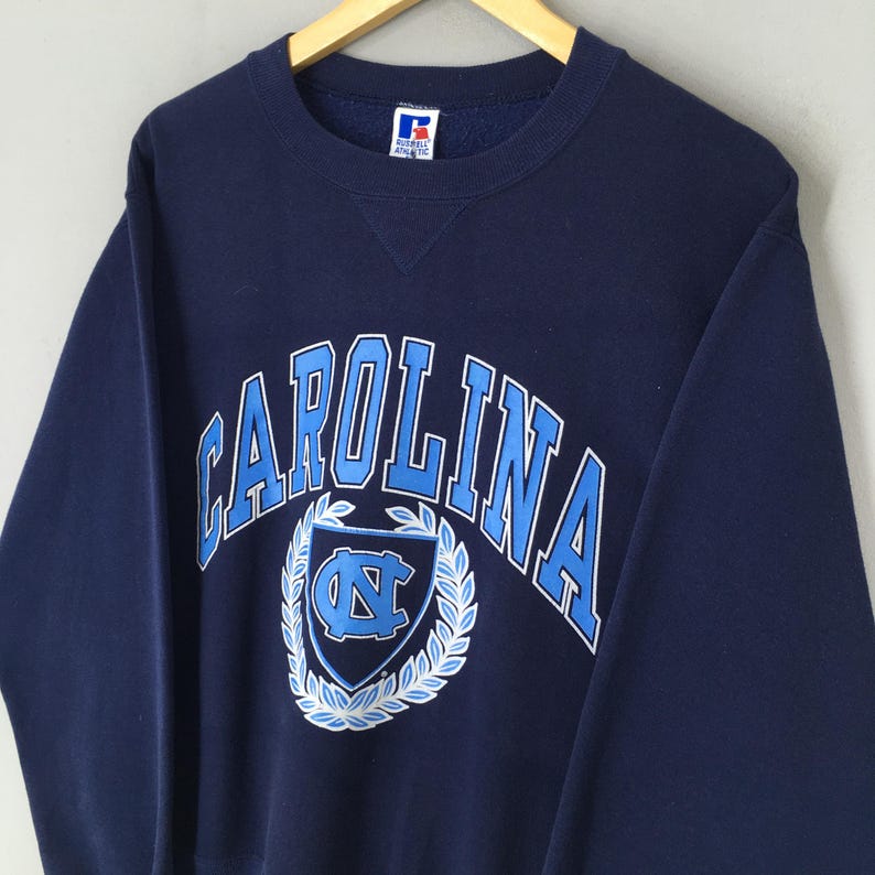 May include: A navy blue sweatshirt with the word "CAROLINA" in light blue block letters. Below the word is a light blue shield with a white wreath and the letters "NC". The sweatshirt has a crew neck and long sleeves.