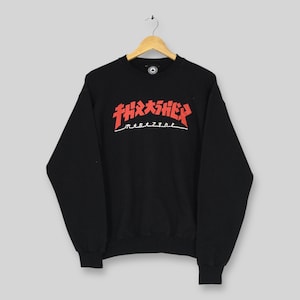 May include: Black crew neck sweatshirt featuring the red "Thrasher Magazine" logo across the chest. The logo is in a stylized font with a white underline. The sweatshirt has long sleeves and ribbed cuffs.
