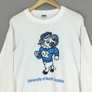 May include: A white long-sleeve crewneck sweatshirt featuring a graphic of the University of North Carolina ram mascot wearing a blue sweater with the letters NC. The text University of North Carolina is printed below in blue, with an NCAA tag inside.