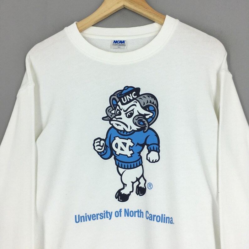 May include: A white long-sleeve crewneck sweatshirt featuring a graphic of the University of North Carolina ram mascot wearing a blue sweater with the letters NC. The text University of North Carolina is printed below in blue, with an NCAA tag inside.
