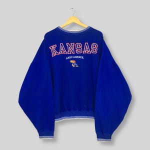 May include: A royal blue sweatshirt with the word "KANSAS" in red block letters across the back. Below the word "KANSAS" is the word "JAYHAWKS" and a cartoon bird. The sweatshirt has white trim around the collar, cuffs, and hem.