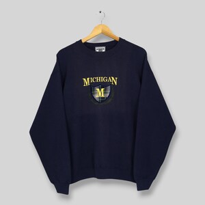 May include: A navy blue crewneck sweatshirt featuring the word MICHIGAN in yellow text above a shield emblem. The shield contains a yellow letter M over a blue and white plaid pattern, surrounded by a thin yellow laurel wreath design.