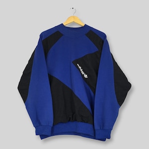 May include: A long-sleeve pullover sweatshirt featuring a color-blocked design in royal blue and black. The garment has a mock neck collar, ribbed cuffs, and a ribbed hem. The Adidas logo and text are embroidered in white on the chest area.