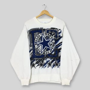 Puede incluir: Sudadera blanca con un gran logotipo de estrella azul y las palabras "Dallas Cowboys" en un diseño circular. El diseño incluye detalles en azul y gris, con cuatro estrellas negras en la parte inferior. Ropa deportiva vintage.