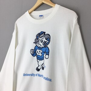 May include: A white long-sleeve crewneck sweatshirt featuring a graphic of the University of North Carolina ram mascot wearing a blue sweater with the NC logo. The text UNC is on the ram's cap, and University of North Carolina is printed below.
