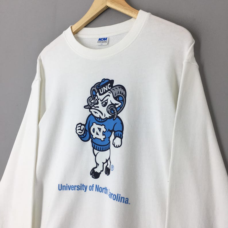 May include: A white long-sleeve crewneck sweatshirt featuring a graphic of the University of North Carolina ram mascot wearing a blue sweater with the NC logo. The text UNC is on the ram's cap, and University of North Carolina is printed below.