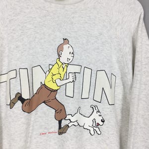 Peut inclure: Un sweat-shirt gris clair avec un dessin de Tintin courant avec Milou. Le mot "TINTIN" est imprim&eacute; en grandes lettres blanches. Tintin porte une chemise jaune et un pantalon marron. Col rond. Design de Moulinsart.