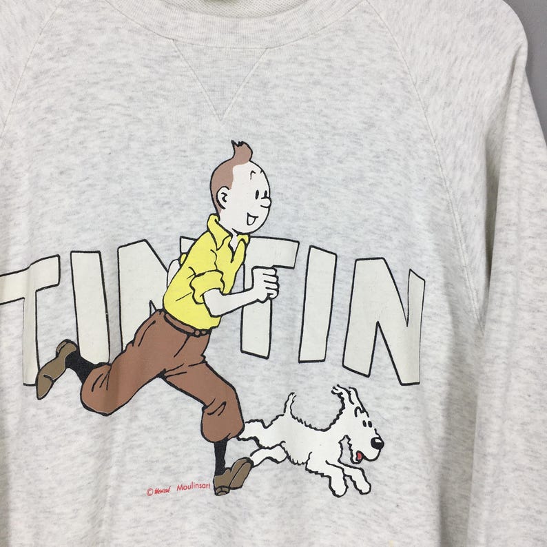 Peut inclure: Un sweat-shirt gris clair avec un dessin de Tintin courant avec Milou. Le mot "TINTIN" est imprim&eacute; en grandes lettres blanches. Tintin porte une chemise jaune et un pantalon marron. Col rond. Design de Moulinsart.