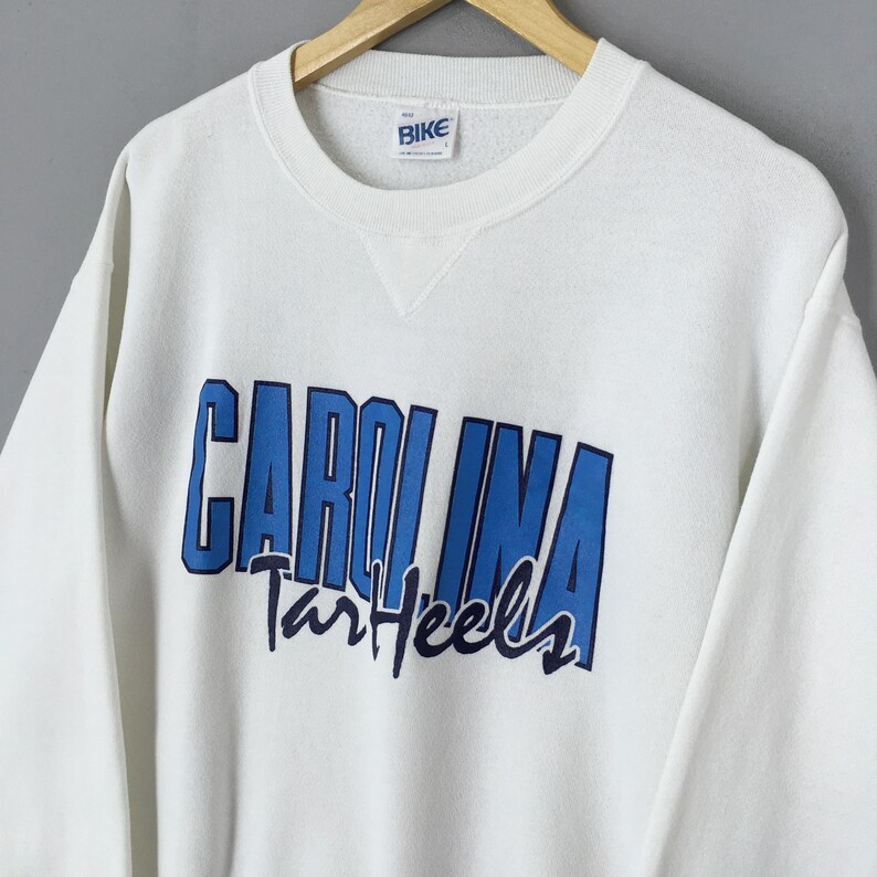May include: White crew neck sweatshirt with the text "CAROLINA Tar Heels" in blue. The sweatshirt has a small "BIKE" tag on the collar. The sweatshirt is hanging on a wooden hanger.