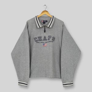 May include: A gray Chaps Ralph Lauren sweatshirt with a quarter-zip collar. The collar and cuffs have white and navy blue stripes. The word "CHAPS" is embroidered across the chest, with a small American flag below.