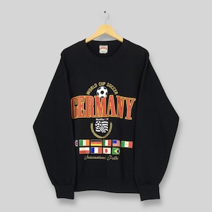 May include: Black sweatshirt with the text "World Cup Soccer Germany" in orange and white. Below the text are flags of various nations and the words "International Pride". The sweatshirt is hanging on a wooden hanger.