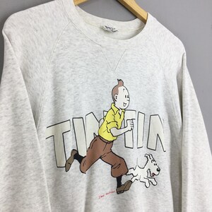 Peut inclure: Sweat-shirt gris clair avec un dessin de Tintin courant avec Milou. Le mot "TINTIN" est imprim&eacute; en blanc. Tintin porte une chemise jaune et un pantalon marron. Le sweat-shirt est suspendu &agrave; un cintre en bois.