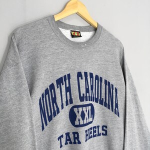 Puede incluir: Una sudadera gris con las palabras "NORTH CAROLINA" en azul marino arqueadas sobre el pecho. Debajo, un &oacute;valo azul marino con "XXL" en su interior. Las palabras "TAR HEELS" est&aacute;n debajo. La sudadera est&aacute; colgada en una percha de madera.