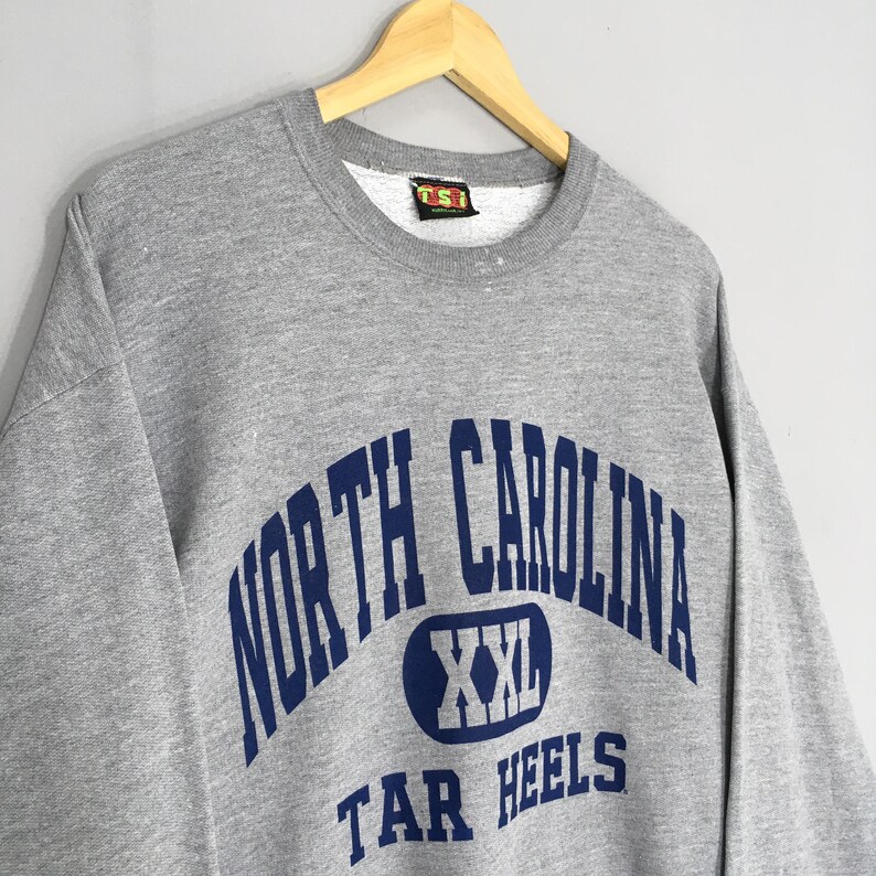 Puede incluir: Una sudadera gris con las palabras "NORTH CAROLINA" en azul marino arqueadas sobre el pecho. Debajo, un &oacute;valo azul marino con "XXL" en su interior. Las palabras "TAR HEELS" est&aacute;n debajo. La sudadera est&aacute; colgada en una percha de madera.